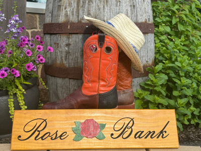 Decorative boot, hat, and wooden sign with 'Rose Bank' text against a rustic background.